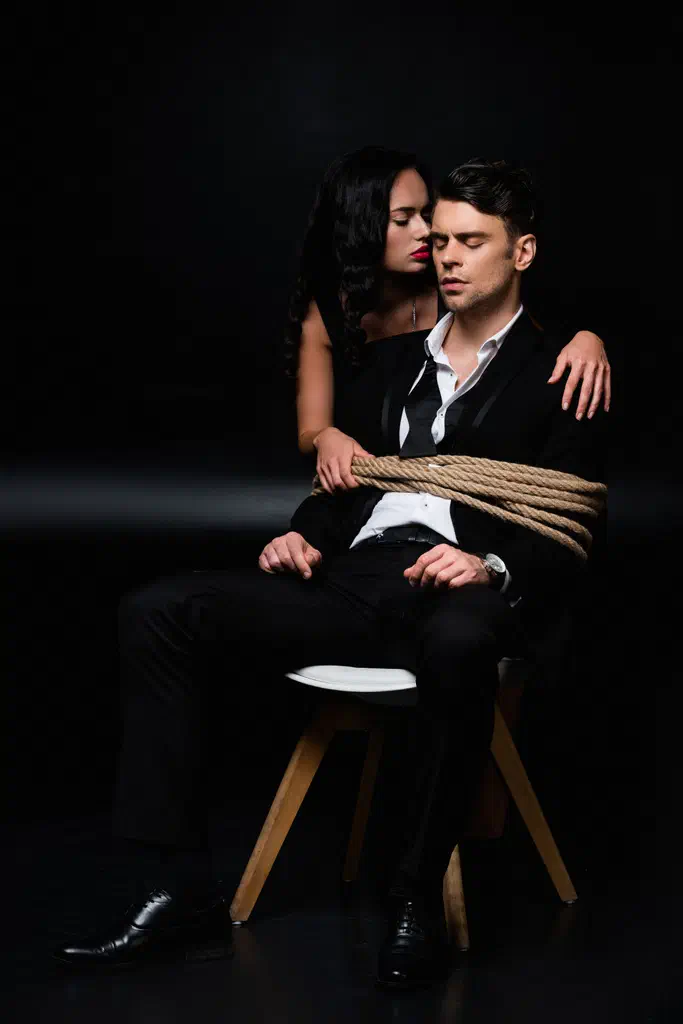 Brunette woman in dress near tied submissive man sitting on chair on black - Stock Photo & Image By VitalikRadko 4912x7360 px